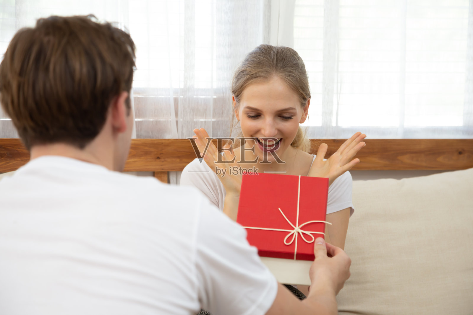年轻男子在起居室的沙发上给漂亮的女友送一个礼盒，惊讶的妻子兴奋而感到惊讶的丈夫，情人节，情侣关系照片摄影图片
