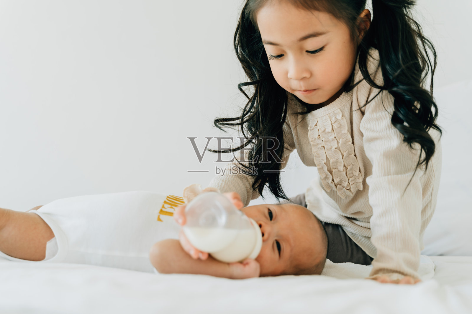 可爱的亚洲家庭小妹妹给刚出生的男婴弟弟喂奶瓶。蹒跚学步的孩子和刚出生的兄弟姐妹在家里的白色卧室里放松照片摄影图片