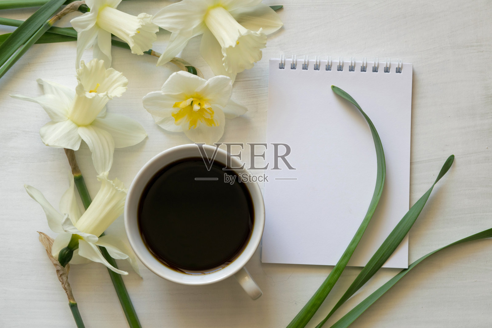 白色桌子上放着记事本、一杯咖啡和水仙花。鼓舞人心的工作场所。照片摄影图片