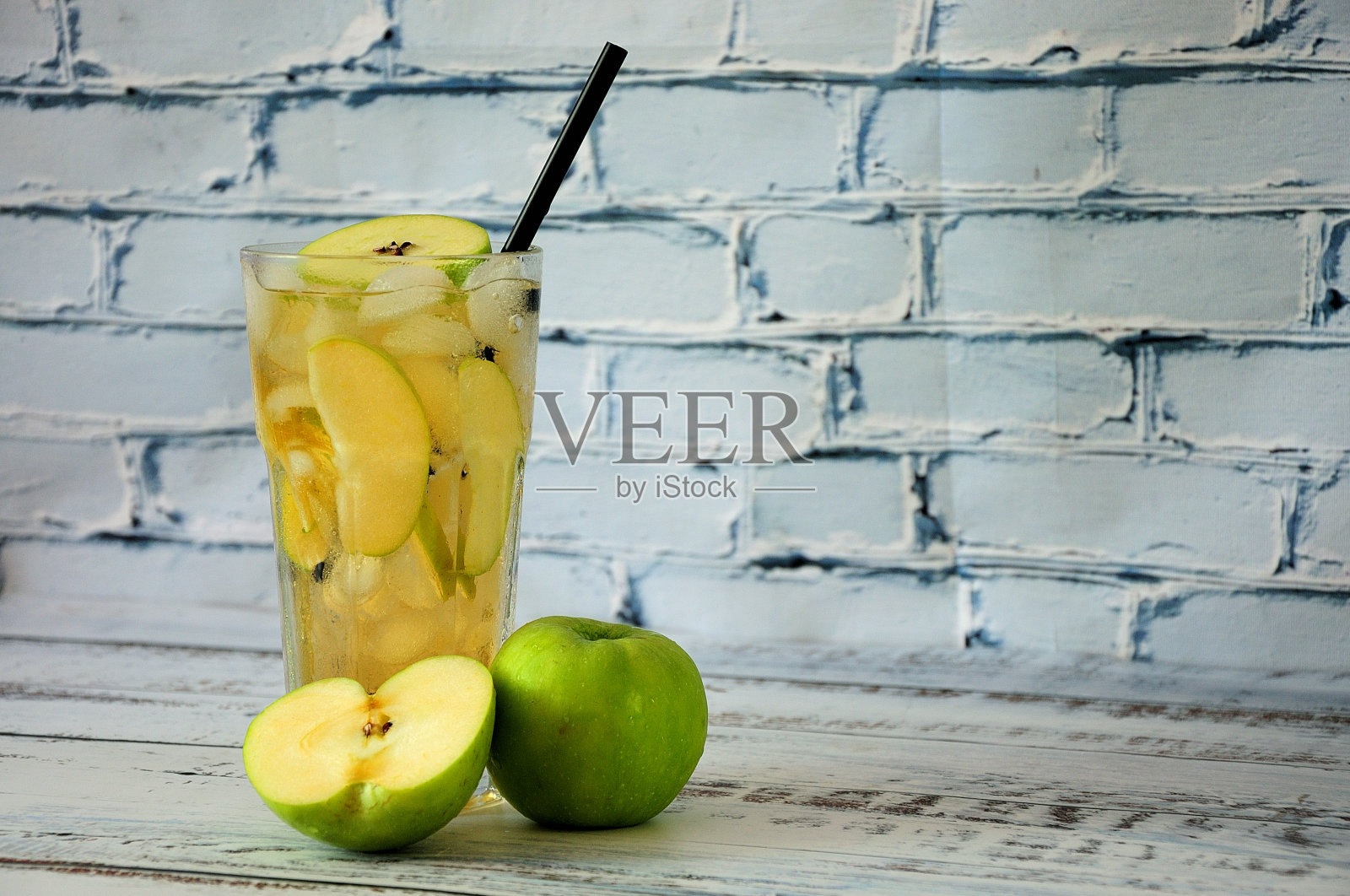 一个装着果汁、冰块和水果片的高脚杯，旁边放着一个切好的绿色苹果，放在砖墙边的木桌上。近距离射杀。照片摄影图片