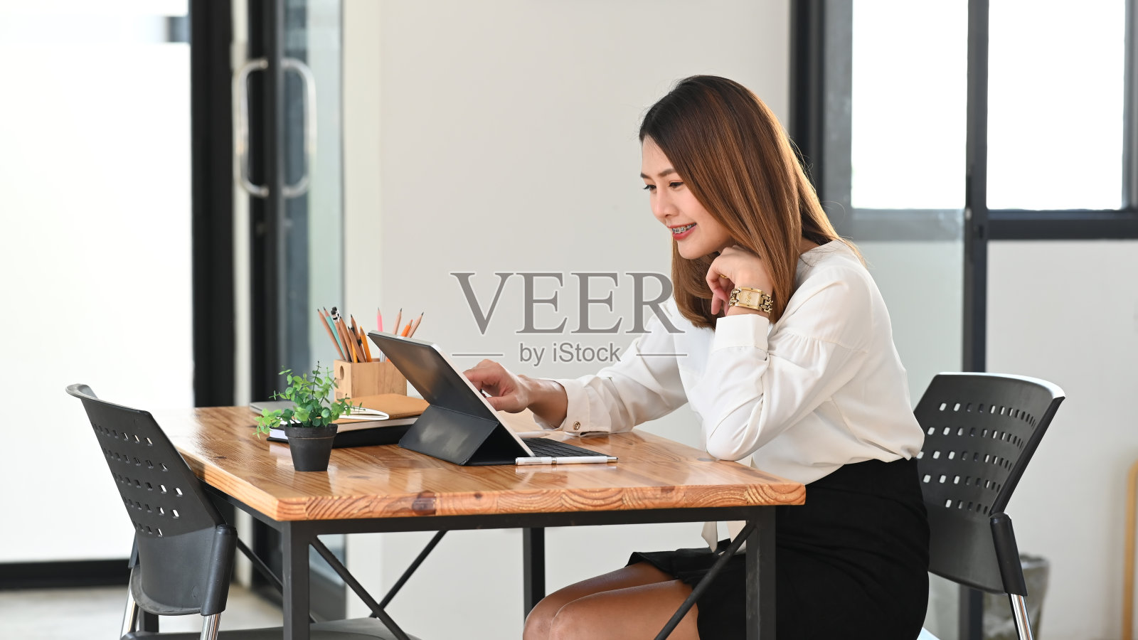 年轻美丽的女子作为秘书使用电脑平板与键盘的情况下，而坐在木制工作桌旁，以有序的办公室为背景。照片摄影图片