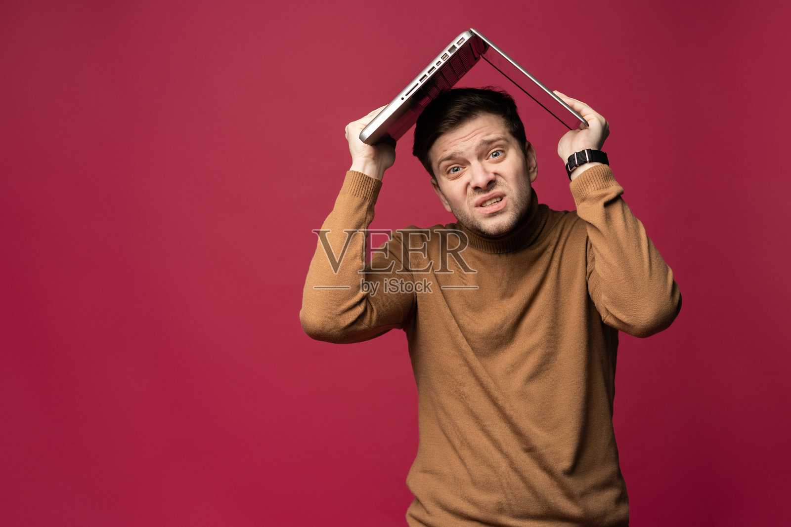 这是一幅不快乐的、紧张的、蓄着胡须的年轻男子的肖像，他把笔记本电脑举过头顶，孤立地站在粉红色的背景下。照片摄影图片