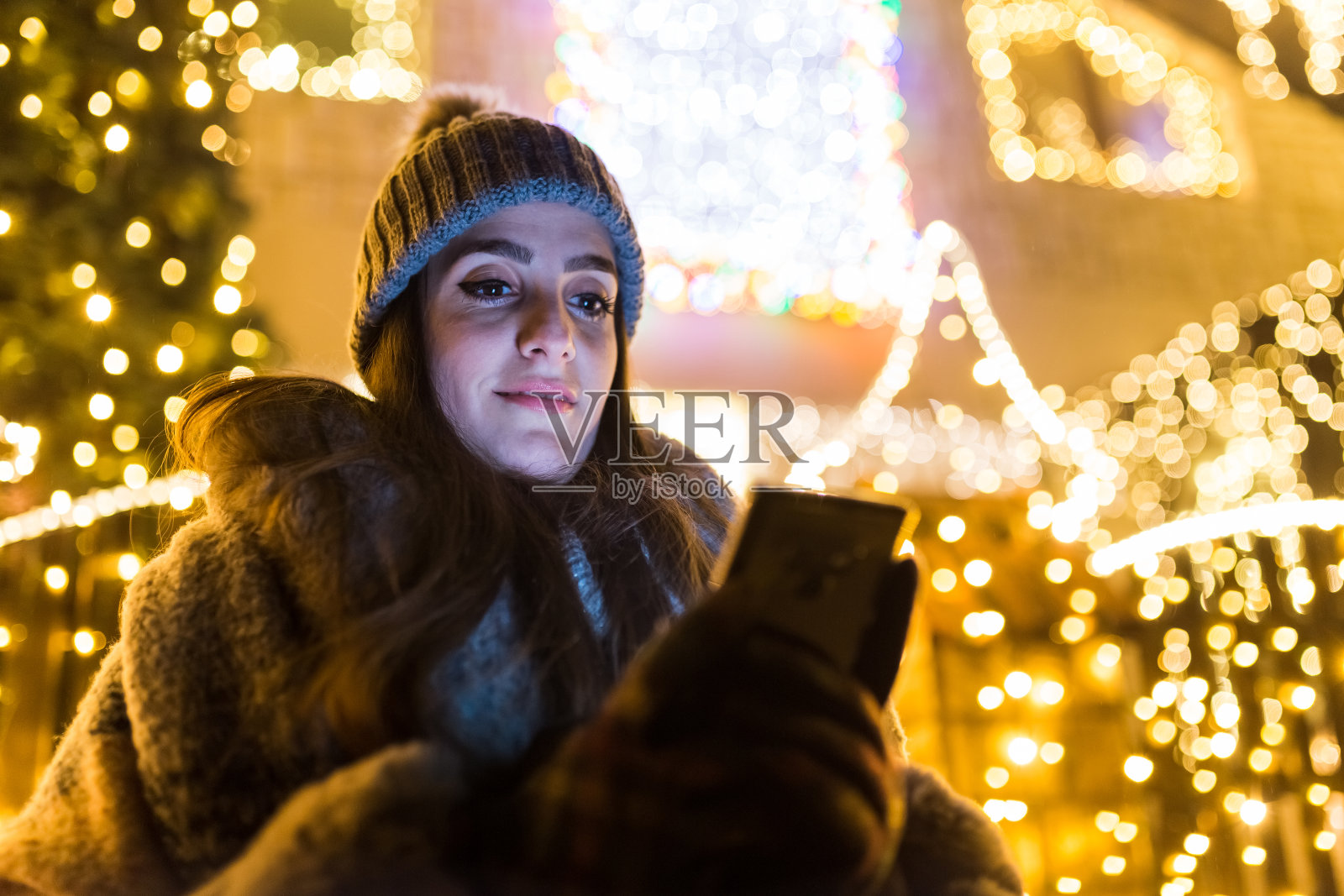美丽的女性拿着手机在用彩灯装饰的房子前。科技与寒假概念。照片摄影图片
