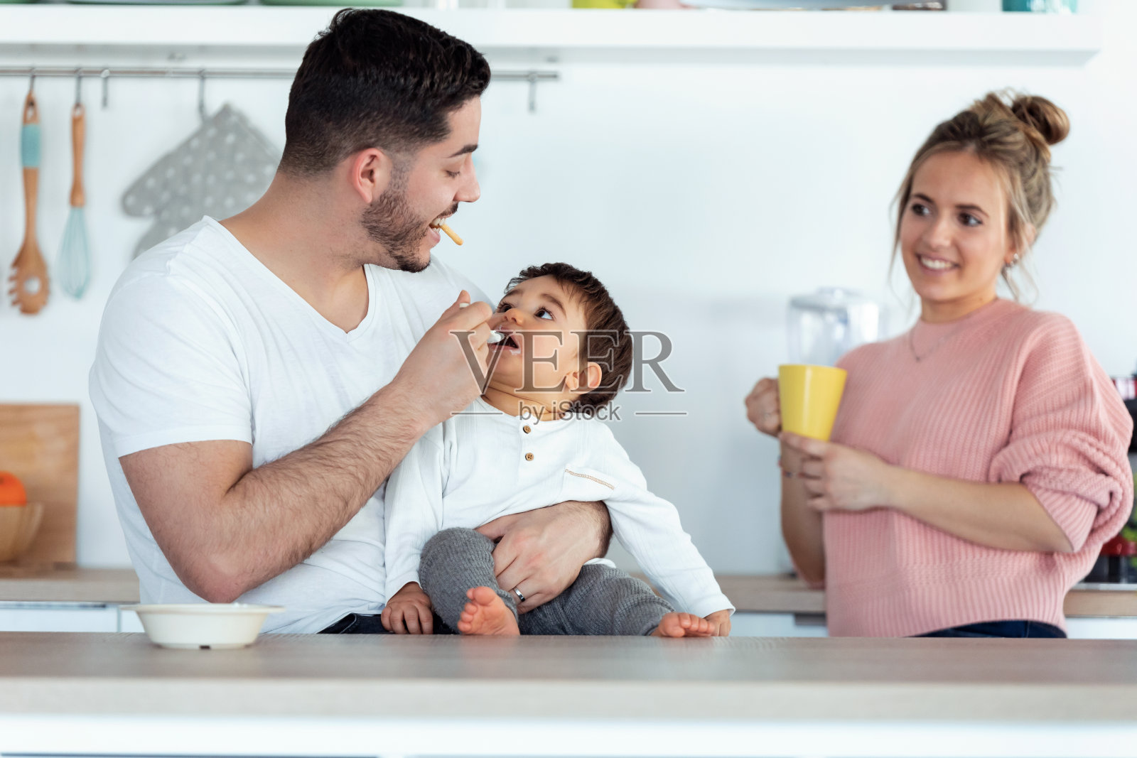 英俊的年轻父亲喂他的婴儿，而母亲看着他们在厨房在家里。照片摄影图片