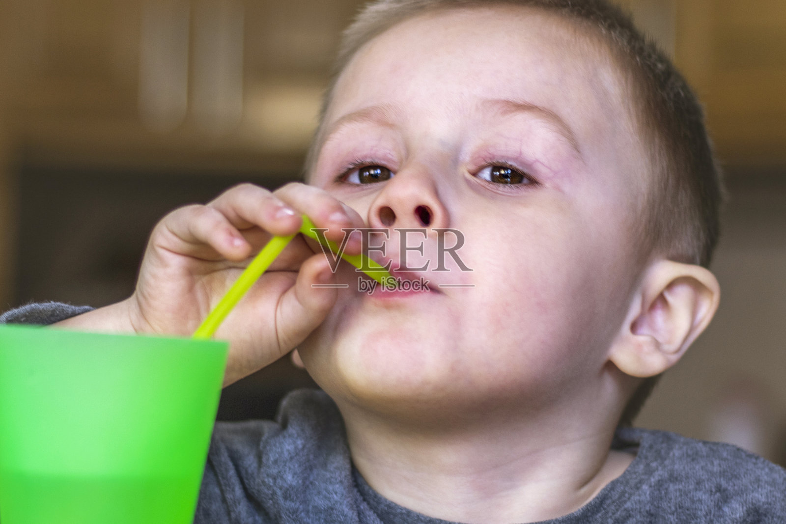 家庭厨房里小孩的特写。漂亮的孩子用吸管喝着绿色塑料杯里的冷饮。照片摄影图片