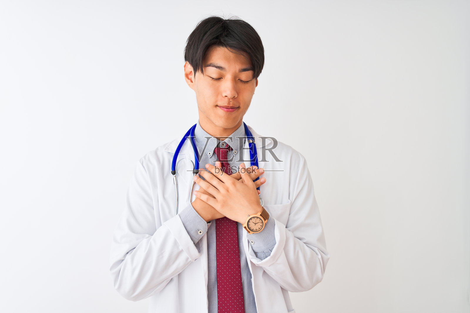 中国医生戴着外套领带，戴着听诊器，在孤立的白色背景下微笑，双手放在胸前，闭着眼睛，脸上做着感激的手势。健康的概念。照片摄影图片
