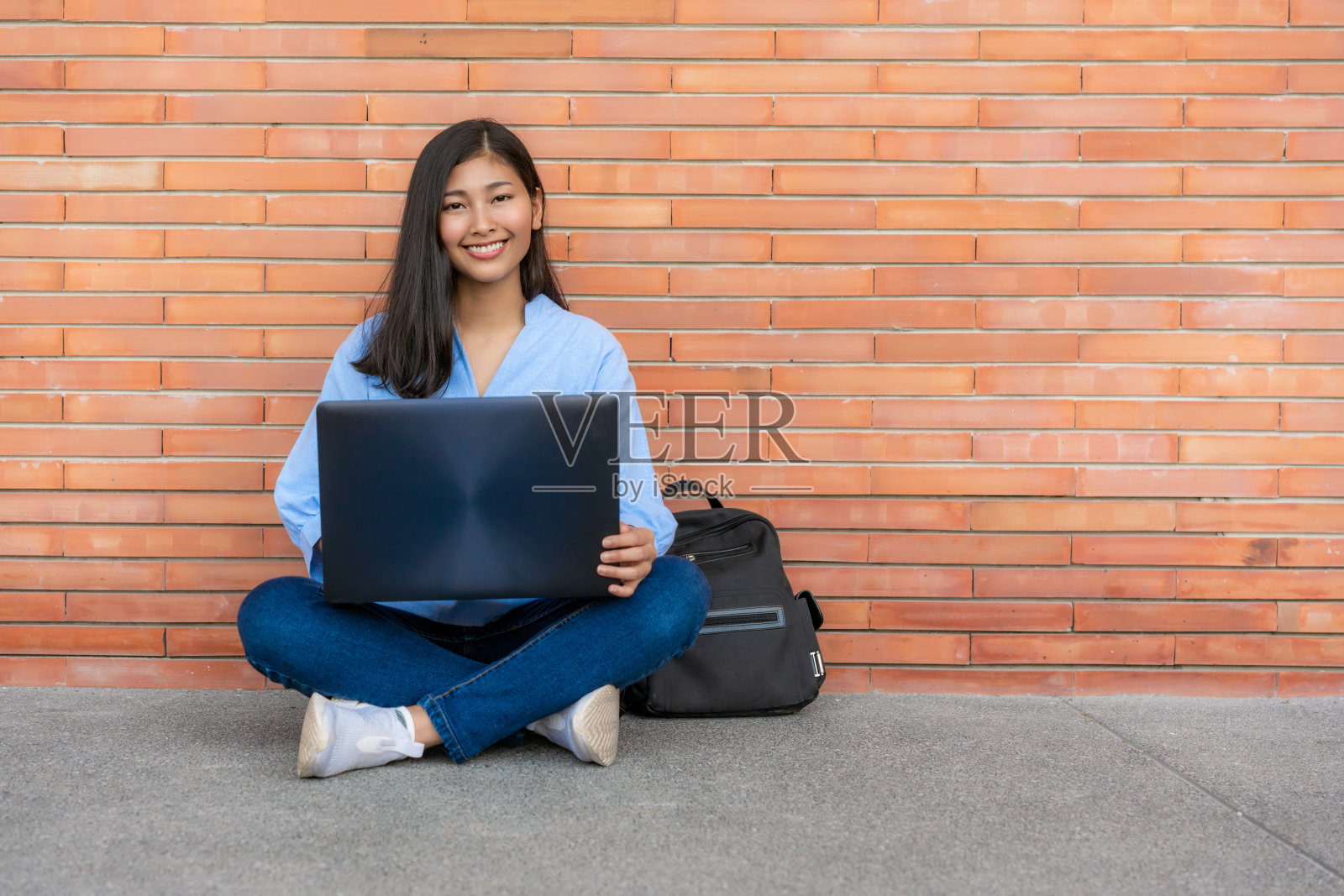 亚洲微笑女学生坐在和使用笔记本电脑在校园砖的背景。快乐的少女高中学生户外。教育，学习，学生，校园，大学，生活理念。照片摄影图片