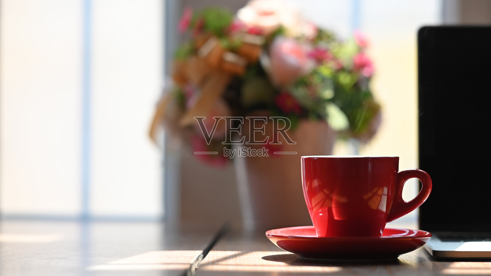 咖啡早晨概念热咖啡红杯杯垫放在木制柜台吧台上，电脑笔记本电脑上模糊的一束花作为背景。照片摄影图片