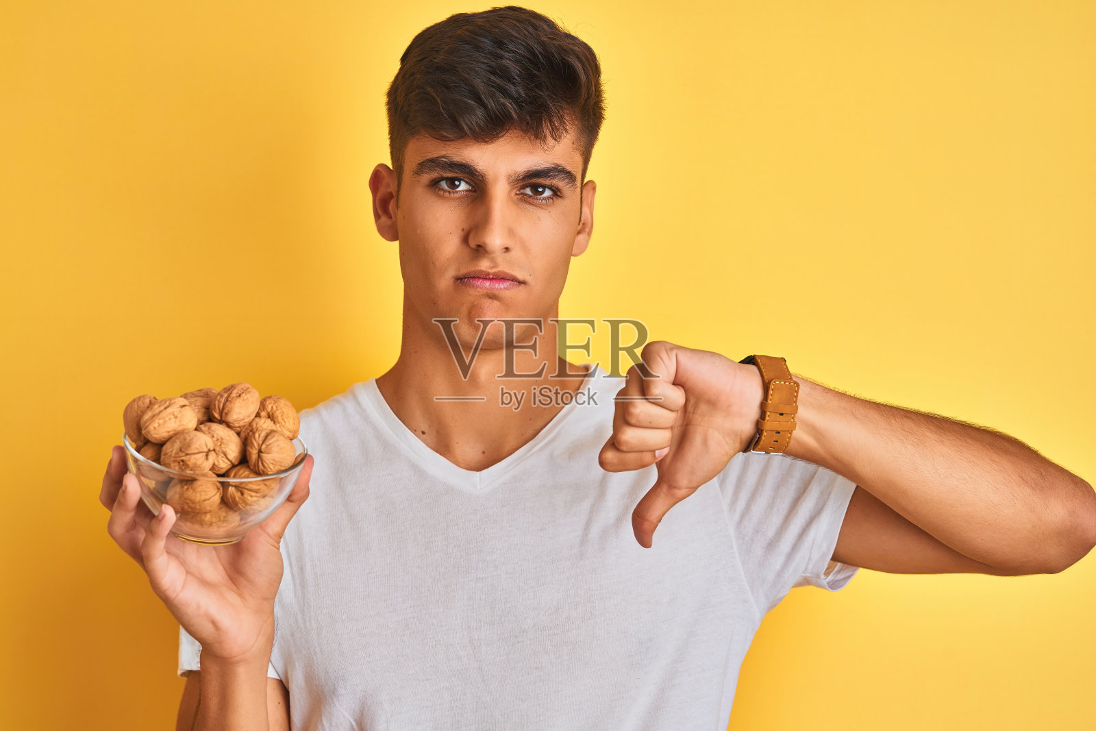年轻的印度男子拿着核桃碗站在孤立的黄色背景与愤怒的脸，消极的迹象显示不喜欢拇指下来，拒绝概念照片摄影图片