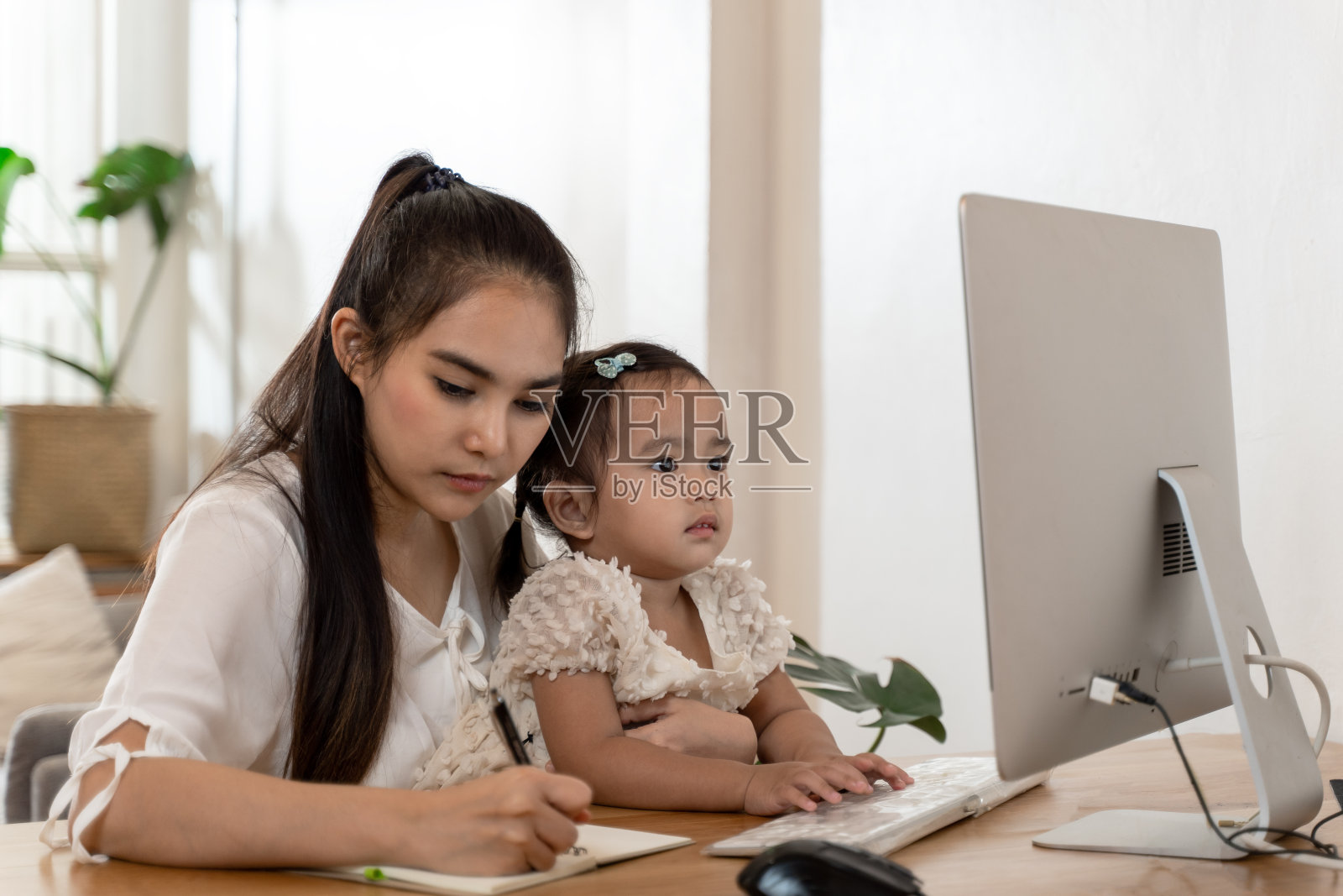 亚洲年轻母亲在家工作，抱着一个孩子，一边讲电话，一边用电脑，一边陪她的孩子照片摄影图片