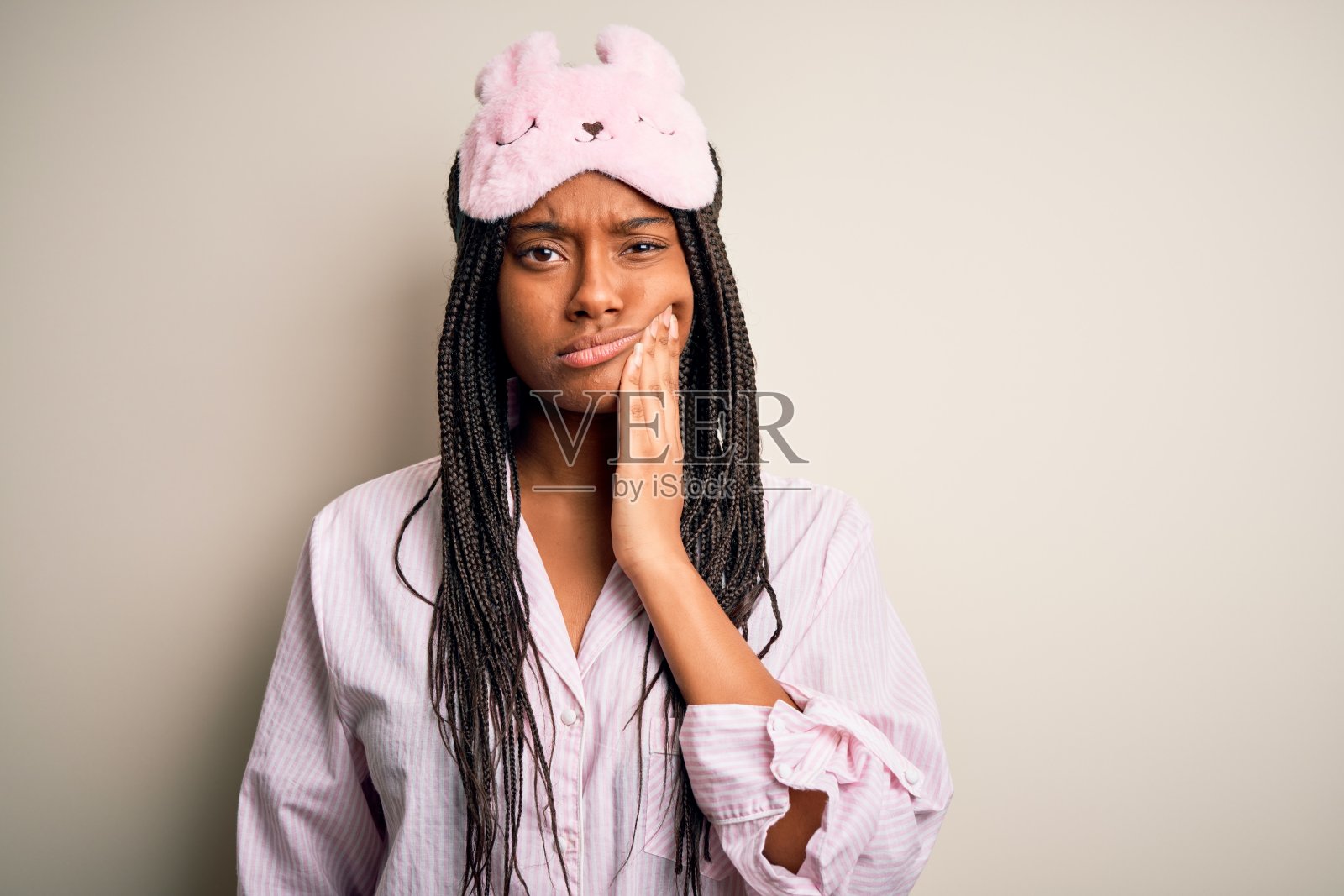 年轻的非洲裔美国妇女，穿着粉红色的睡衣，戴着睡眠面罩，在孤立的背景下用手触摸嘴巴，表情痛苦，因为牙痛或牙齿疾病。牙医照片摄影图片