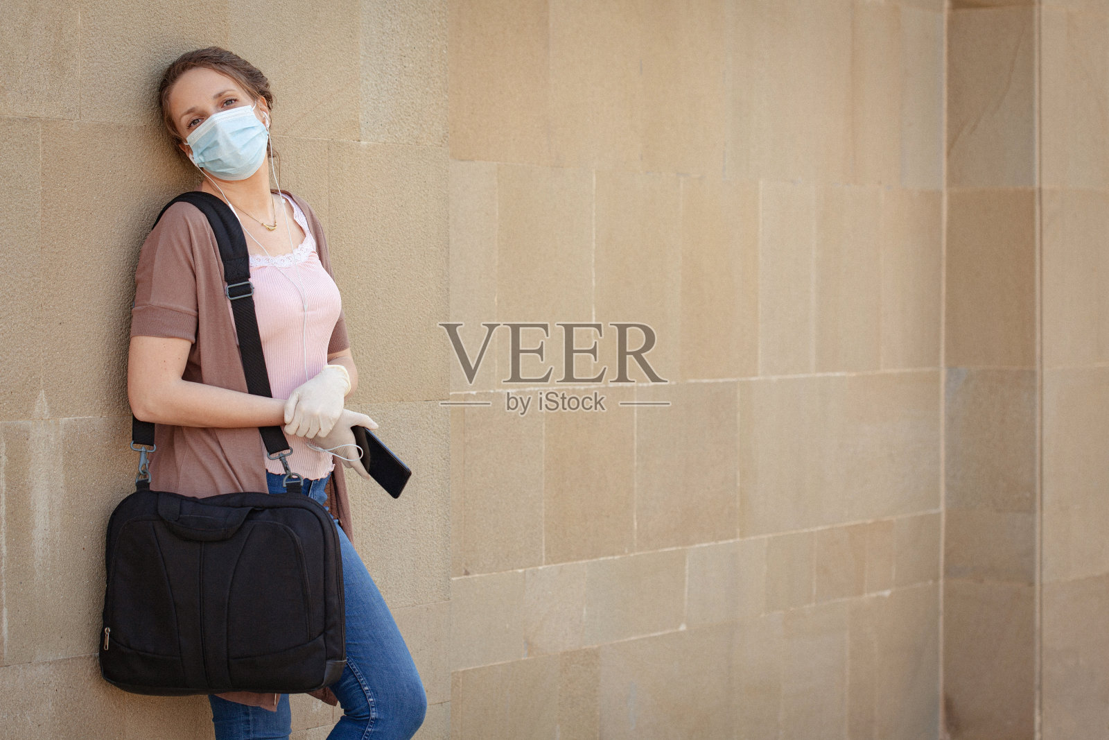 2019冠状病毒病大流行期间，忧心忡忡的妇女戴着防护口罩和手套，靠在石墙上放松照片摄影图片