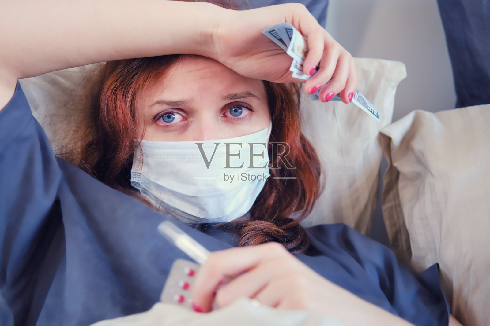 一名染有冠状病毒的红发妇女戴着医用口罩躺在床上，手里拿着美元用于治疗照片摄影图片