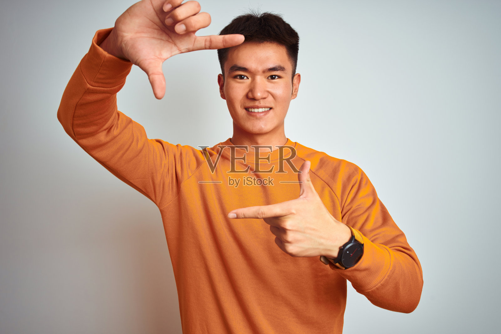 年轻的亚洲华人男子穿着橙色毛衣站在孤立的白色背景微笑与手和手指框架与快乐的脸。创意和摄影概念。照片摄影图片