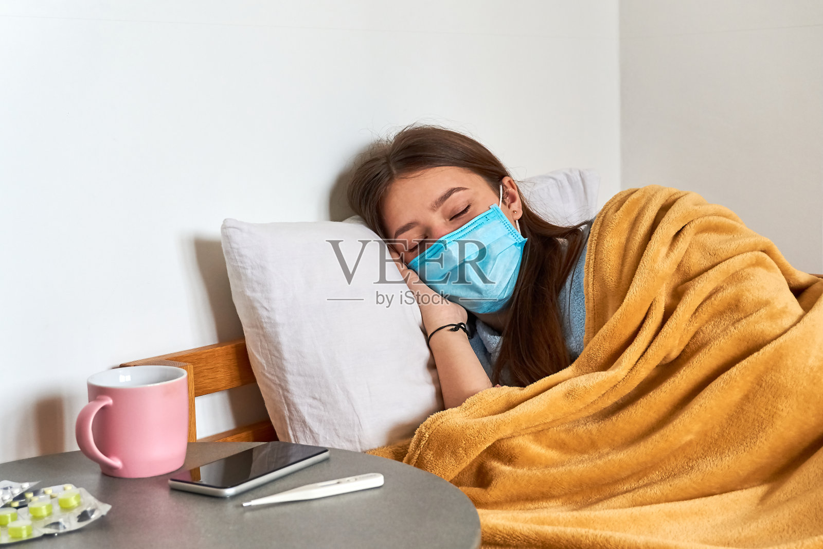 患流感的女孩戴着防护面罩躺在家里的床上。药片、温度计和药片放在桌上。照片摄影图片