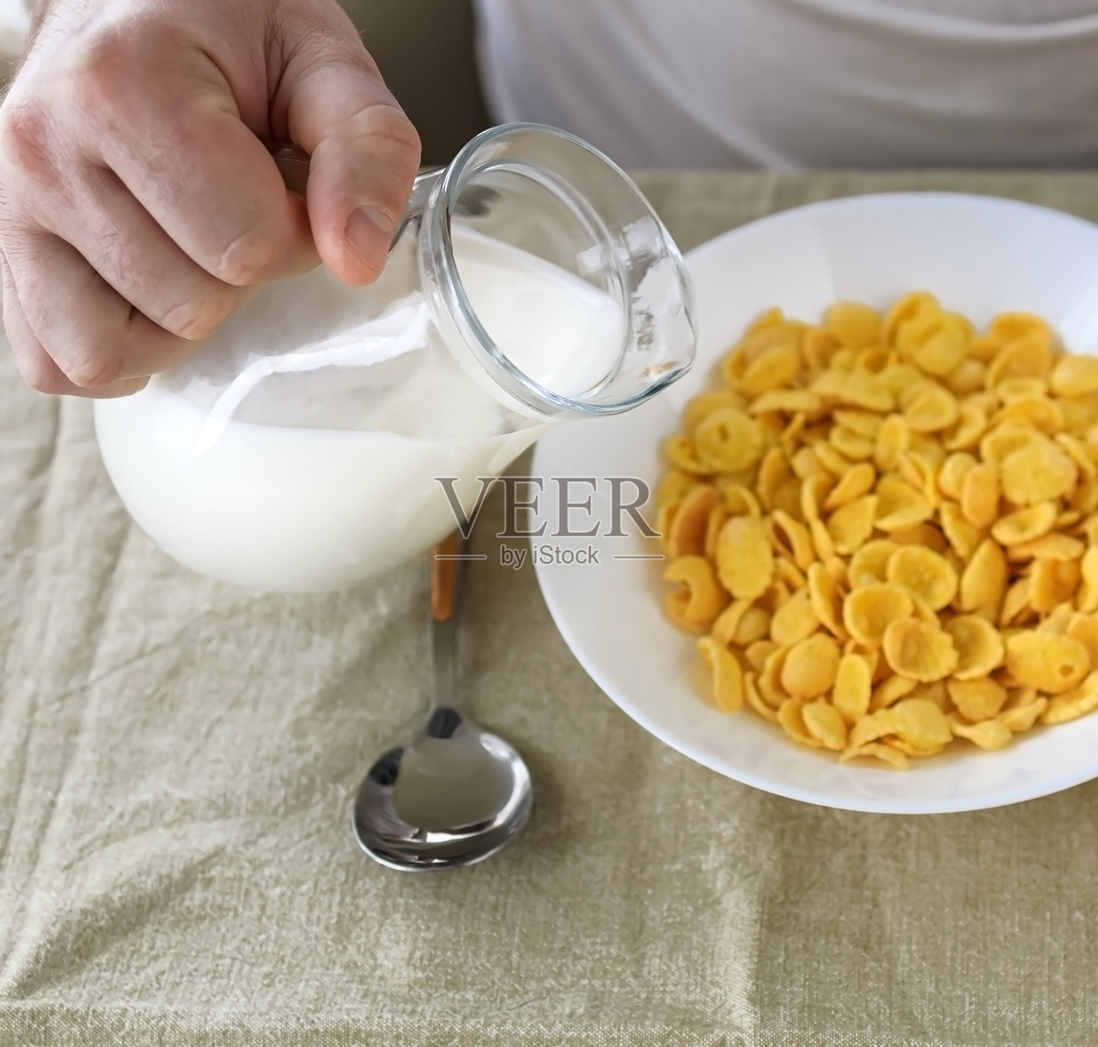 一个身材矮小的男子把牛奶倒进一块朴素粗糙的桌布上的白色盘子里的玉米片里。从以上观点。有选择性的重点。概念，简单快速的美式健康早餐照片摄影图片