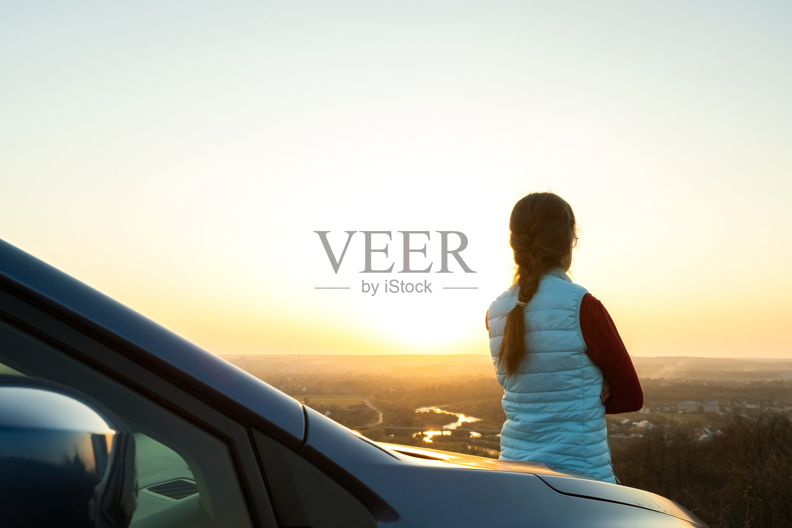 年轻女子站在她的车旁享受温暖的日落景色。女旅行者靠在汽车引擎盖上看着黄昏的地平线。照片摄影图片