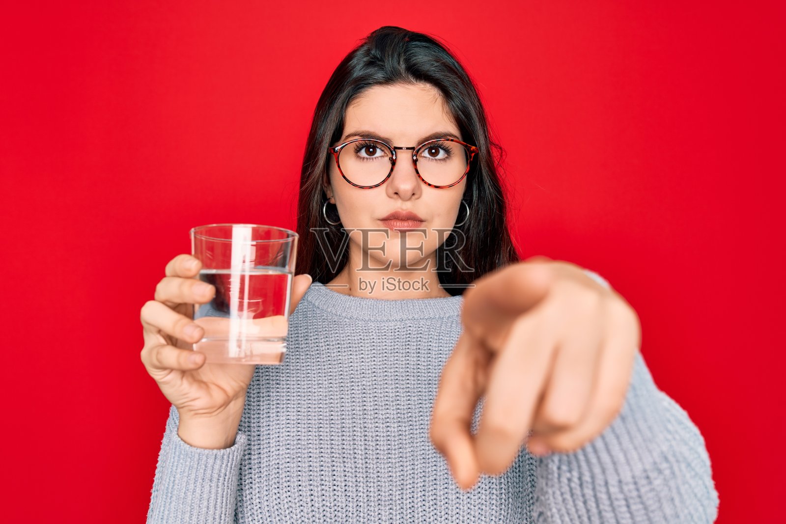 年轻美丽的女人喝一杯新鲜的水在孤立的红色背景指向指向相机和你，手势，积极和自信的手势从前面照片摄影图片