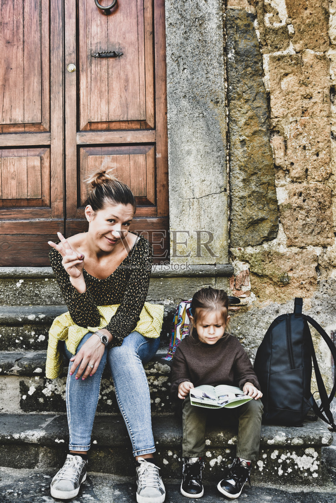 一个女人和她的女儿坐在古老的意大利村庄的木门前的楼梯上。目标达成了，胜利就用手签。小女孩在纸地图中寻找。旅行的概念。照片摄影图片