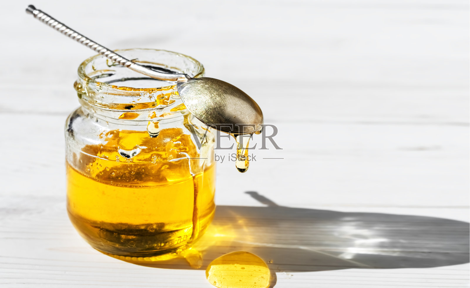 有机天然蜂蜜在一个玻璃罐中，在一个白色的木制背景，特写。老式的勺子放在一罐开着的蜂蜜上，蜂蜜滴在桌上。明亮的阳光透过一罐蜂蜜照射进来。照片摄影图片