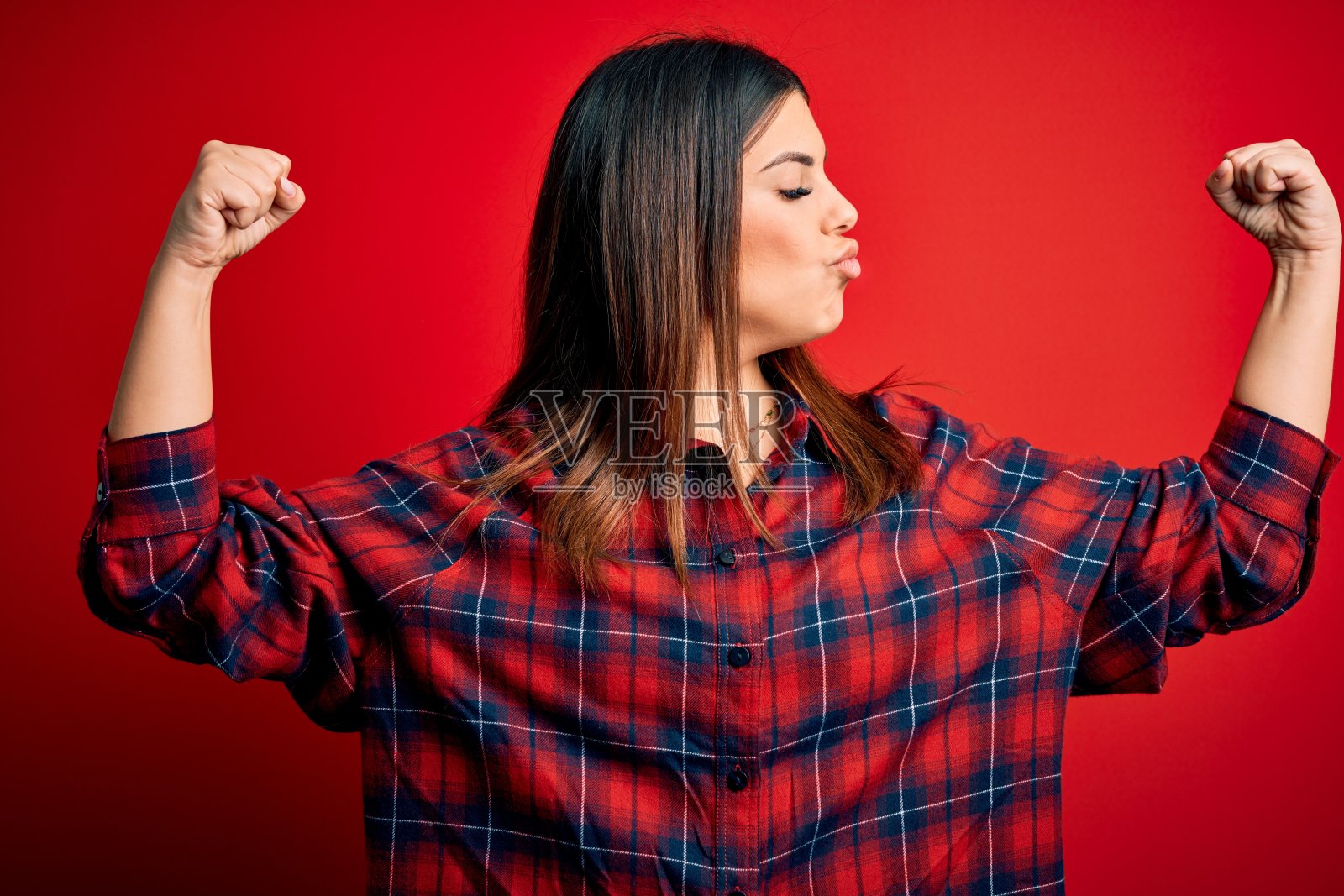 年轻美丽的女子穿着休闲衬衫在红色背景显示手臂肌肉微笑自豪。健康的概念。照片摄影图片