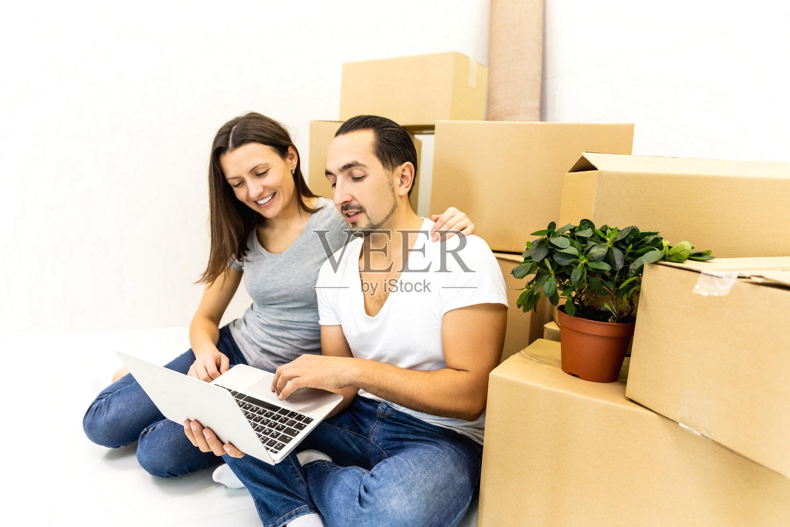 年轻的家庭坐在纸板中间，用笔记本电脑上网寻找家具到他们的新房子。照片摄影图片