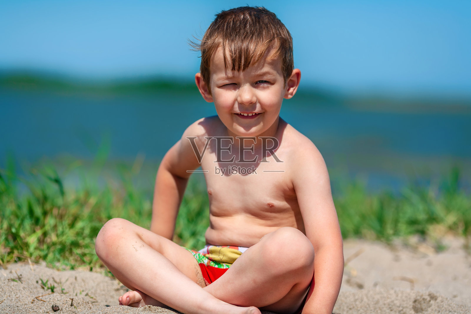中午，一个穿着短裤的小男孩赤裸着上身坐在湖上的沙滩上。照片摄影图片