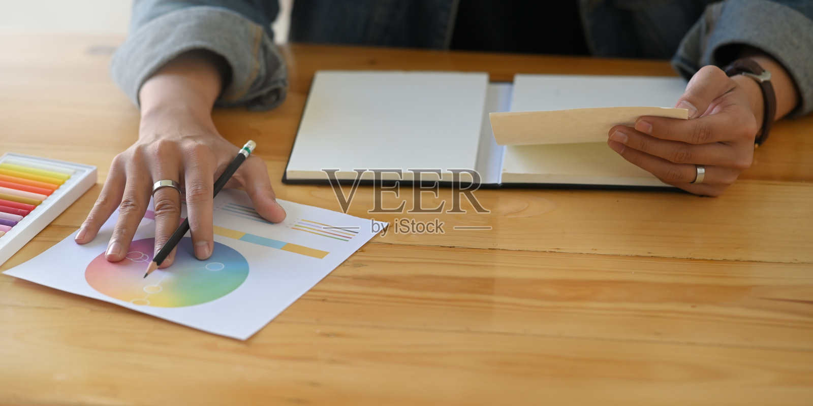 裁剪图像的设计师的手拿着一支铅笔和纸，而坐在前蜡笔，颜色指南和颜色表在木制办公桌上舒适的工作室作为背景。照片摄影图片
