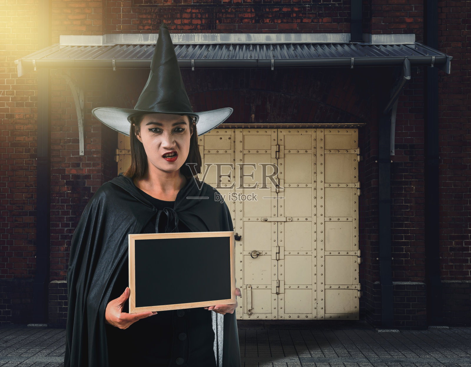 一个穿着黑色吓人的女巫万圣节服装的女人，拿着写有女巫古老城堡老砖墙的粉笔板照片摄影图片