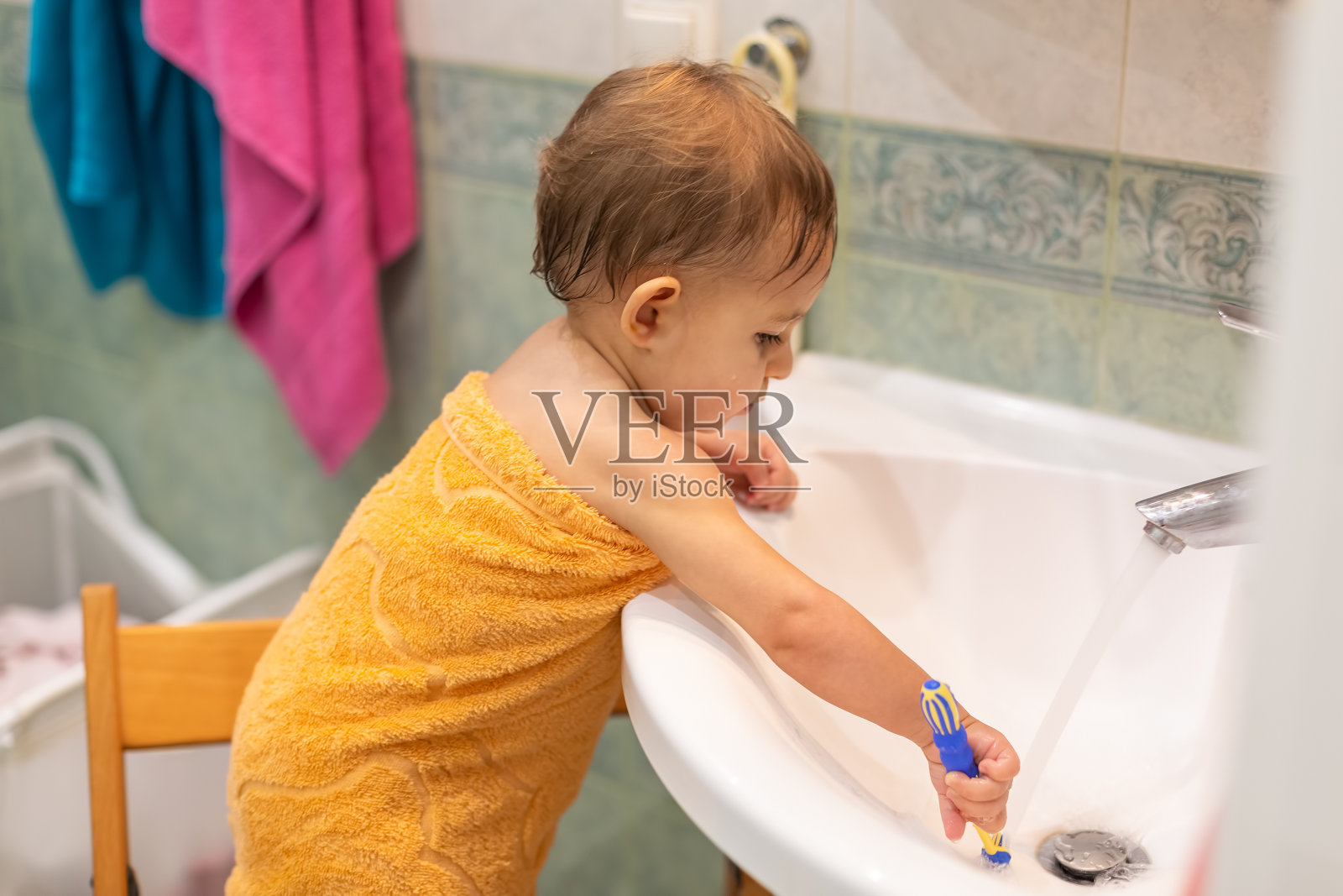 一个1岁的小可爱，站在浴室的椅子上，用橙色的毛巾包裹着牙刷，在水龙头下的水槽里用水清洗。特写，软焦点，模糊背景照片摄影图片