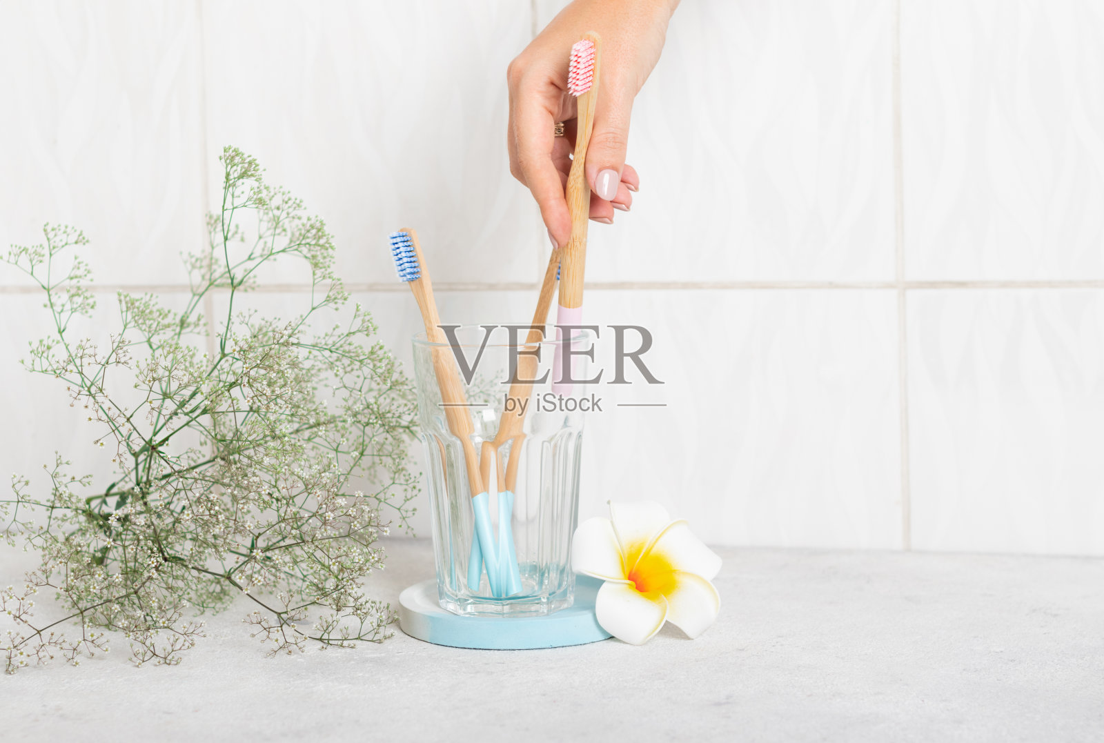 浴室里的玻璃杯子里装着崭新的粉色和蓝色木制竹制牙刷。早上，女性的手拿起一支粉蓝相间的牙刷。可生物降解的个人护理产品。没有塑料的概念。Plumeria照片摄影图片