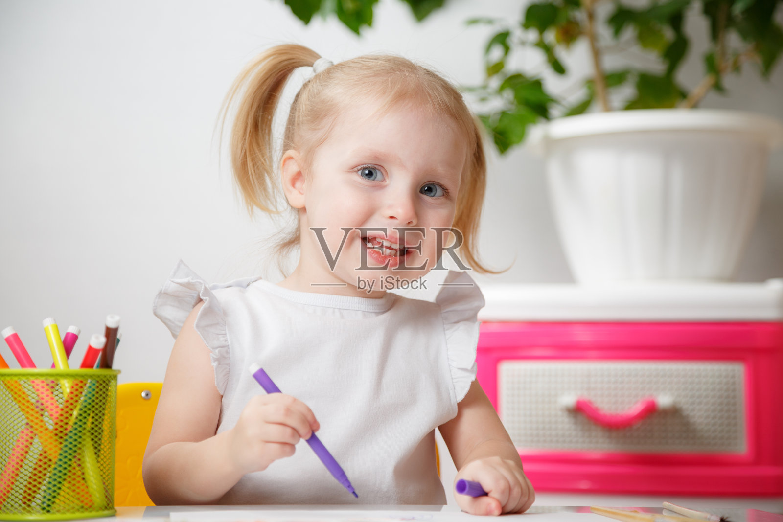 小女孩在家里或幼儿园的桌子上画画。可爱可爱的小孩子与两个马尾辫绘制室内。彩色记号笔盒子。照片摄影图片