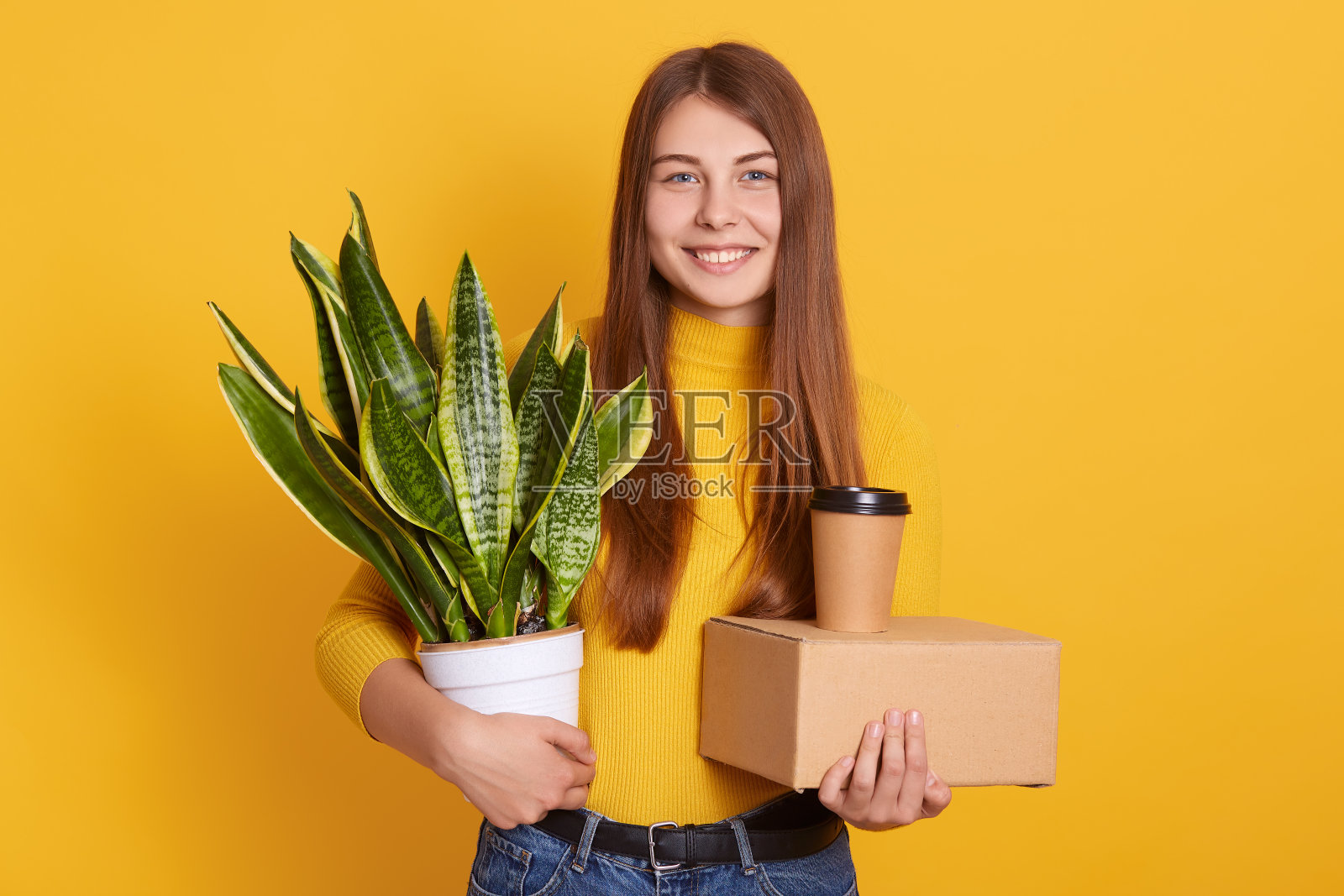 快乐好看的女孩穿着休闲服装孤立地站在黄色的演播室背景，看着微笑的相机，搬迁，移动到其他，手持盒子，外卖咖啡和植物。照片摄影图片