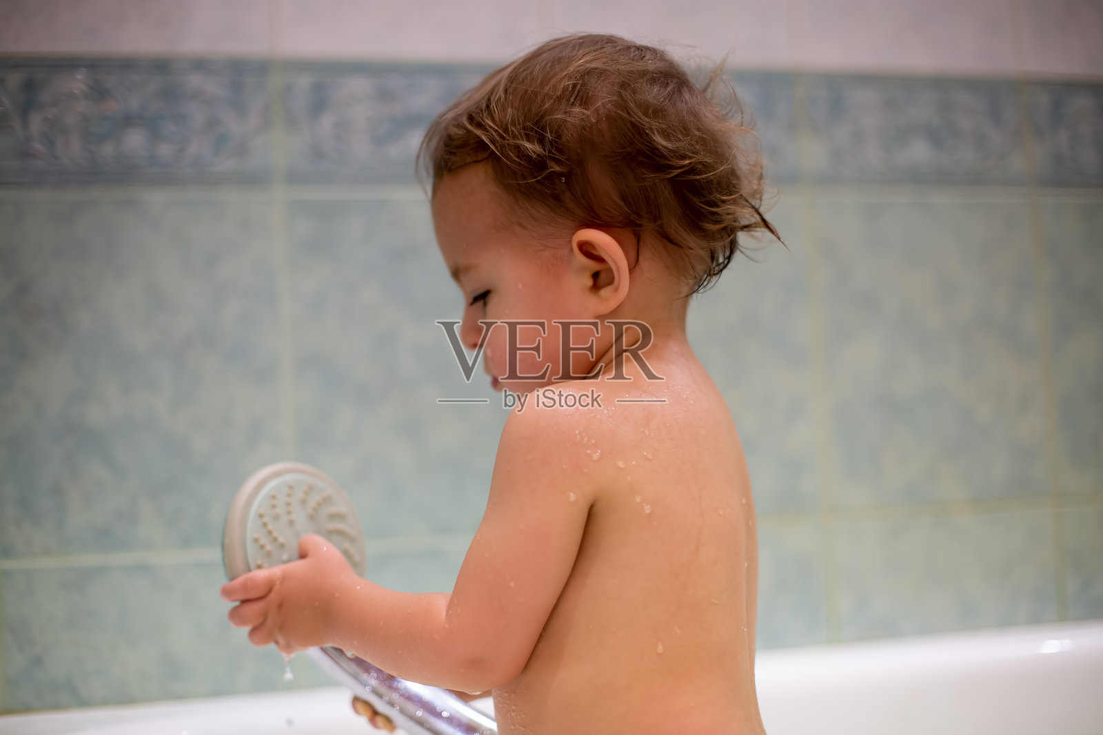 一个可爱的小婴儿站在浴缸里洗澡，手里拿着喷壶。灵魂在玩弄它。以绿色浴室为背景，模糊的特写，柔和的焦点照片摄影图片