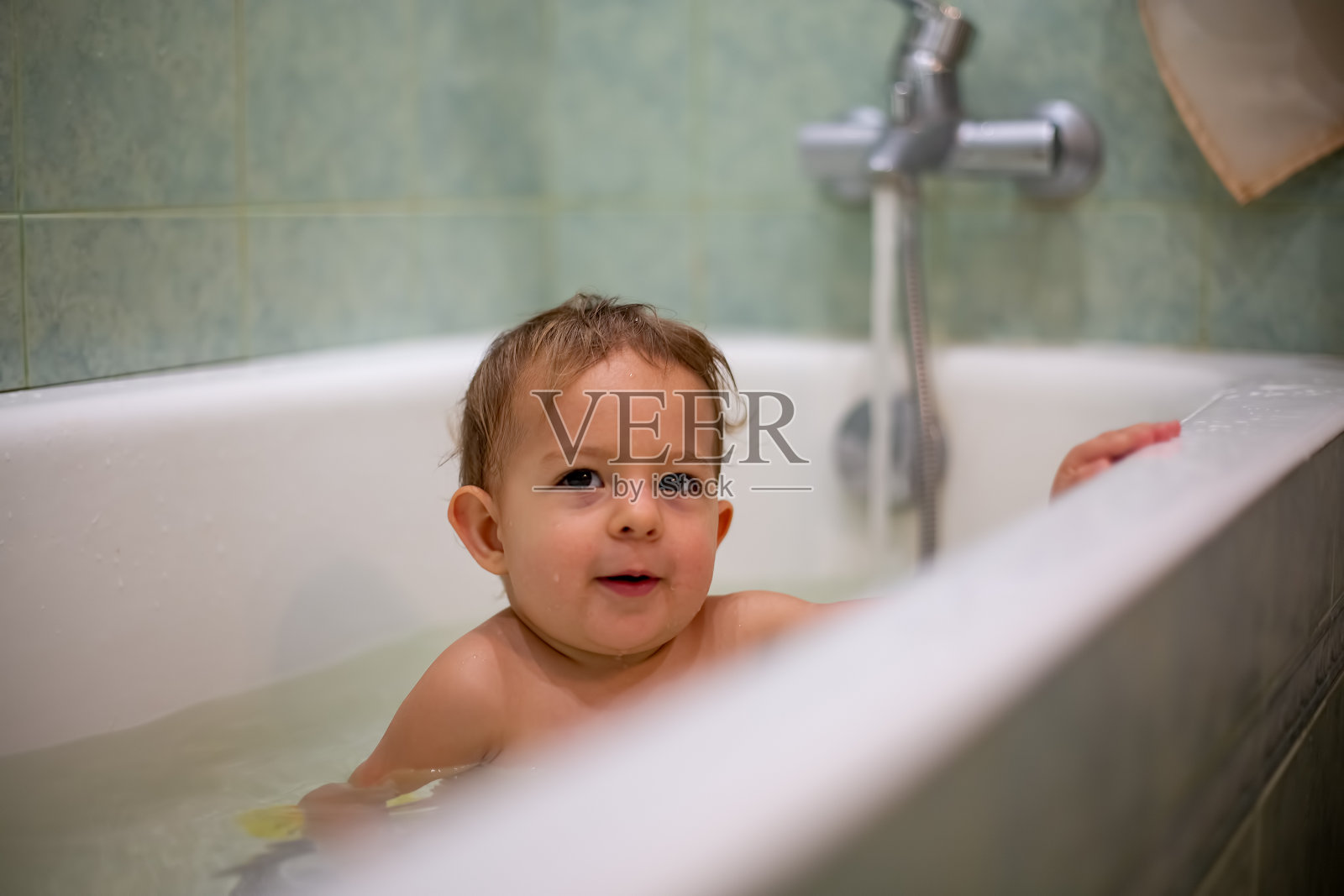 可爱的白人婴儿在家里洗澡，偷偷地微笑着休息在浴缸的一边。水飞溅，背景是一间模糊的绿色浴室。特写,软焦点照片摄影图片
