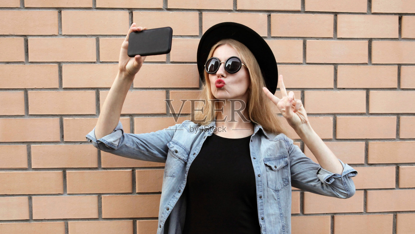 美丽的年轻女子用智能手机自拍，吹红色的嘴唇发送甜蜜的飞吻戴着黑色圆帽，牛仔裤夹克在砖墙的背景照片摄影图片