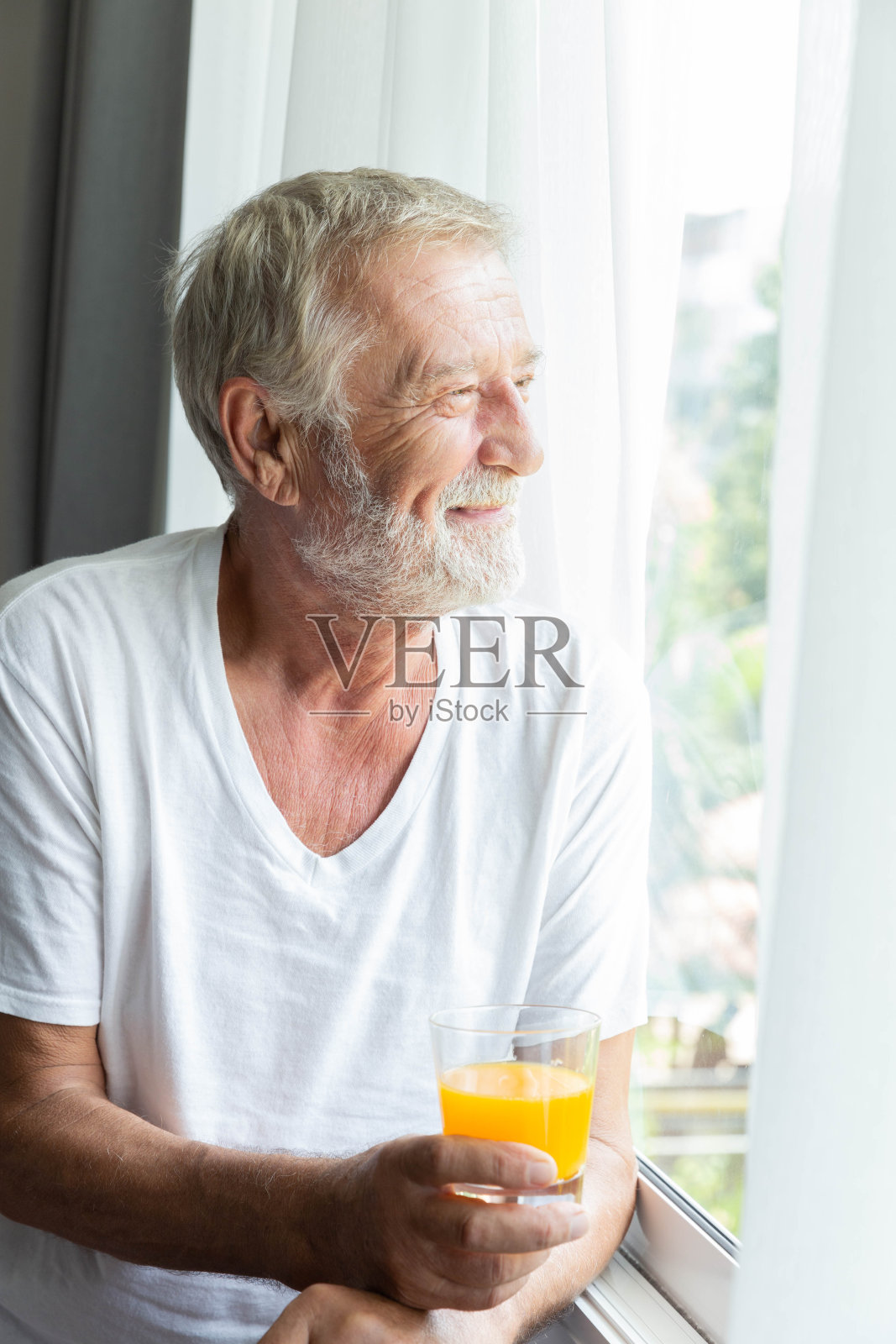 一位退休老人站在房间窗户旁，手里拿着一杯橙汁，幸福地望着外面照片摄影图片