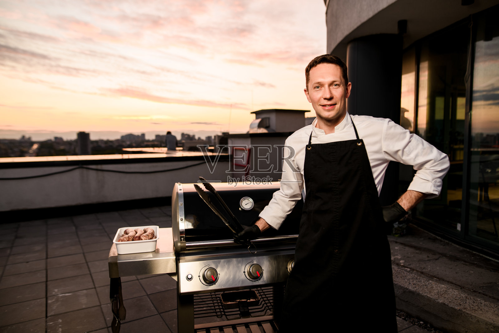 面带微笑的男厨师手里拿着火钳，站在烧烤设备旁边。照片摄影图片