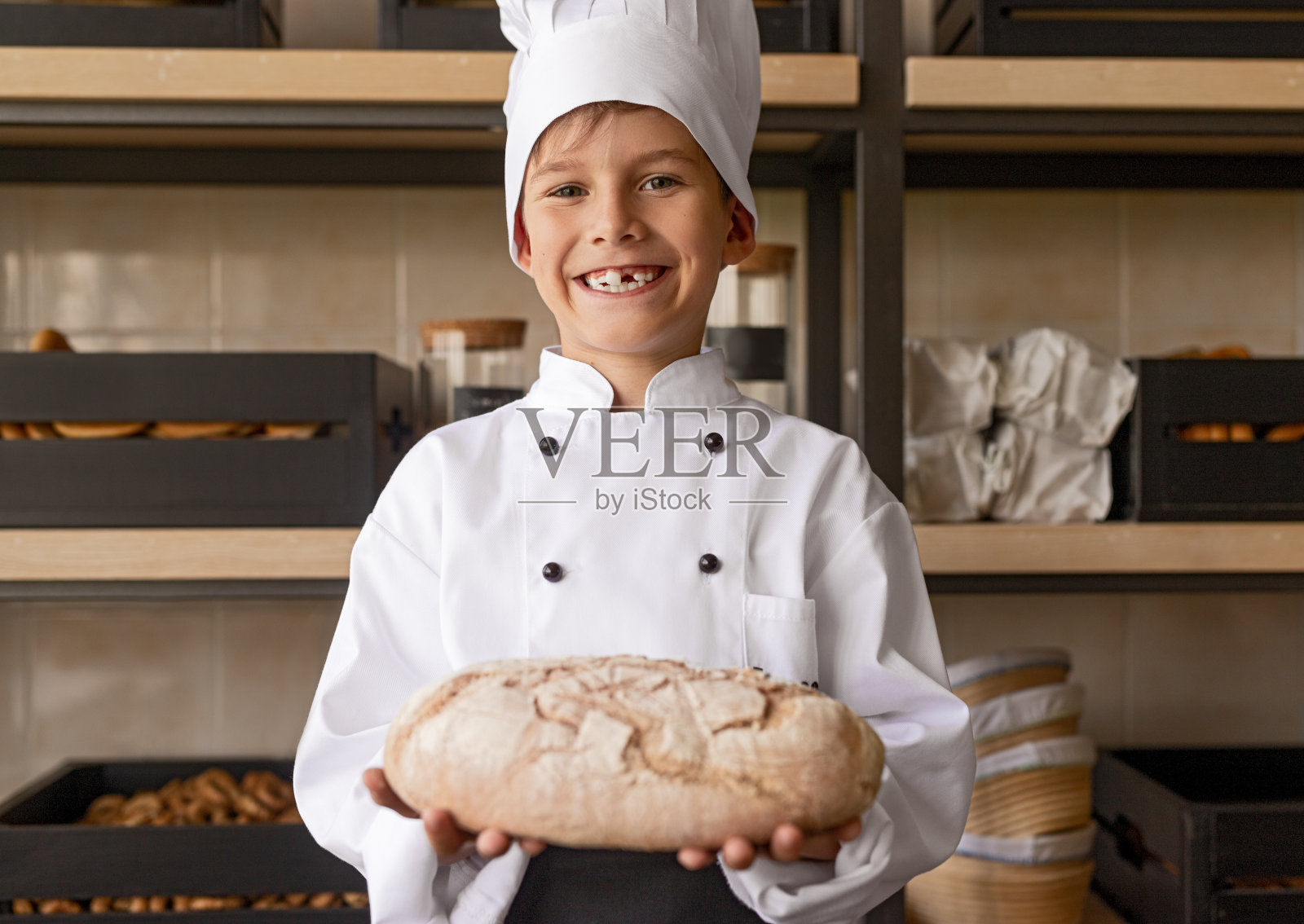 骄傲的小面包师穿着制服拿着新鲜的糕点站在面包店照片摄影图片