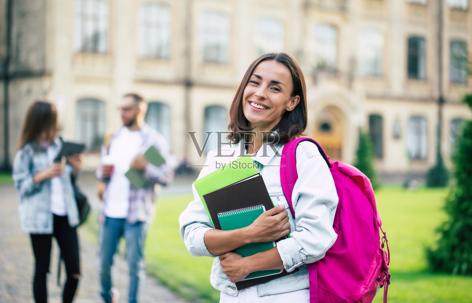 年轻漂亮的黑发学生女孩穿着牛仔衣服，背着背包，手里拿着书，看着镜头上的一群她的学生朋友的背景照片摄影图片