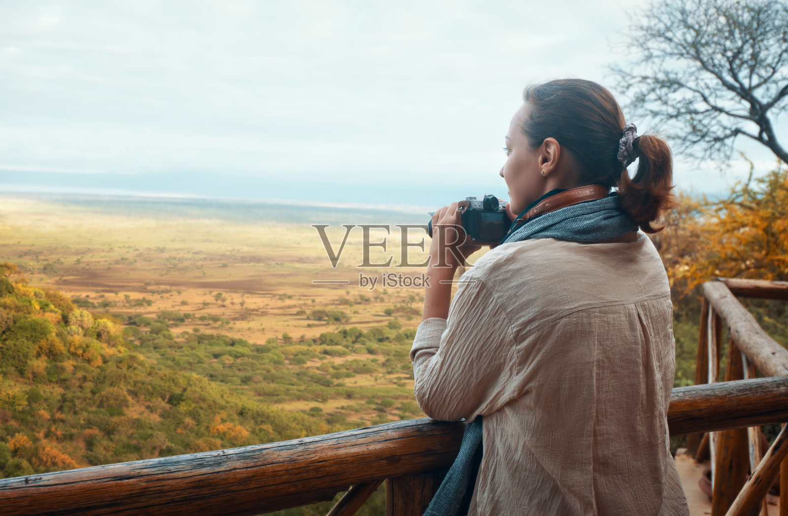 一名女子站在木质的阳台上，拿着相机看着非洲大草原。照片摄影图片
