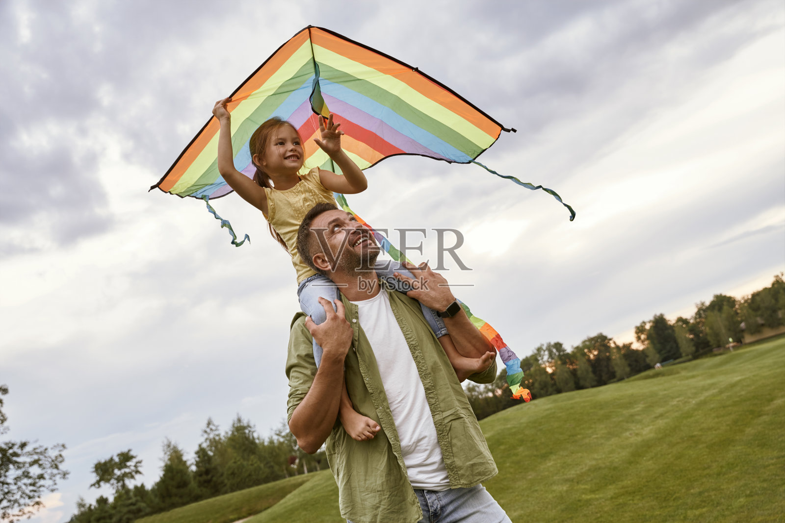 可爱的微笑小女孩坐在她的爸爸的肩膀上玩五颜六色的风筝，小女儿和爸爸一起在公园里度过一个温暖的日子照片摄影图片