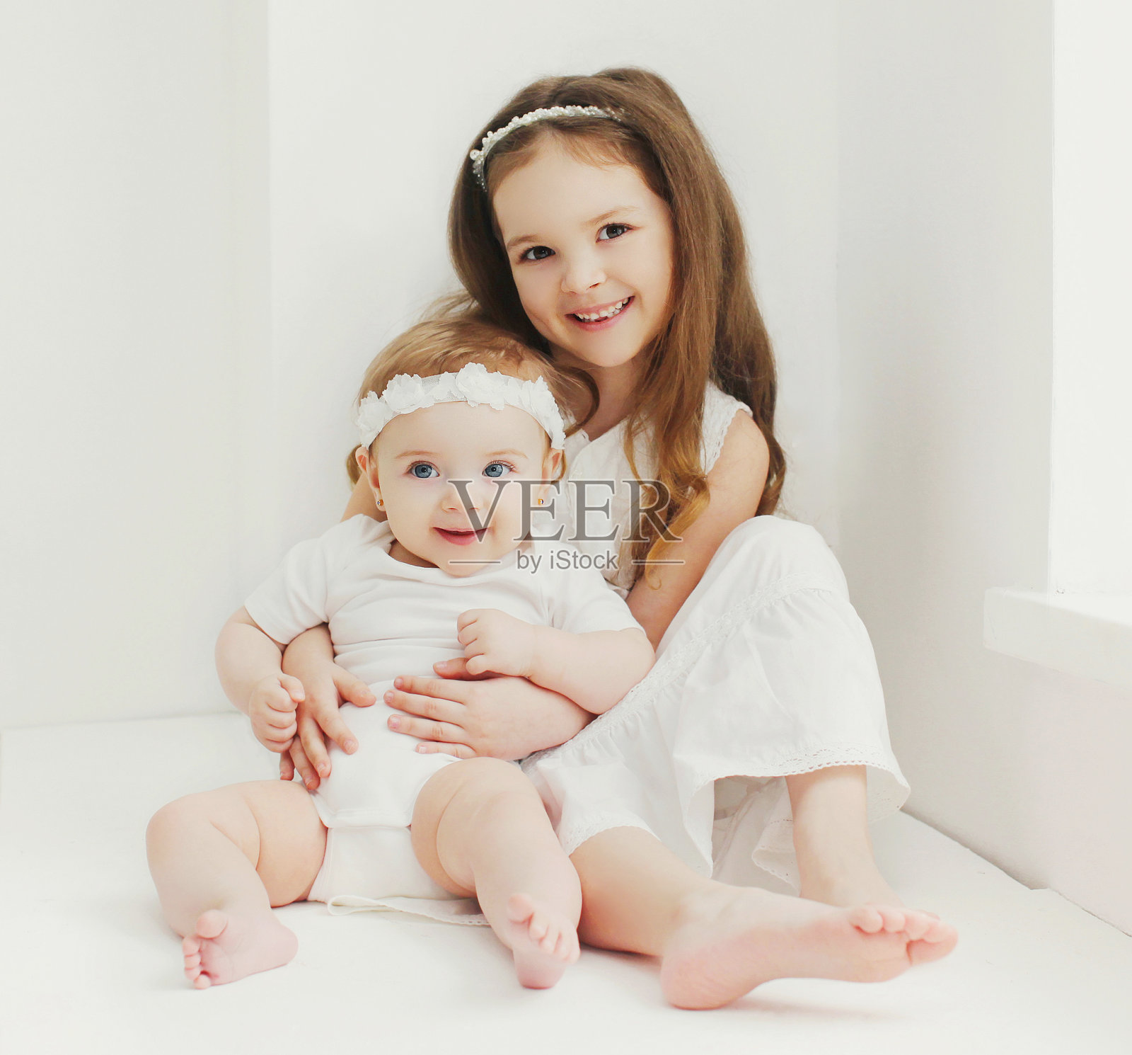 可爱的小女孩和她的妹妹婴儿坐在家里靠近窗户的白色房间里的肖像照片摄影图片