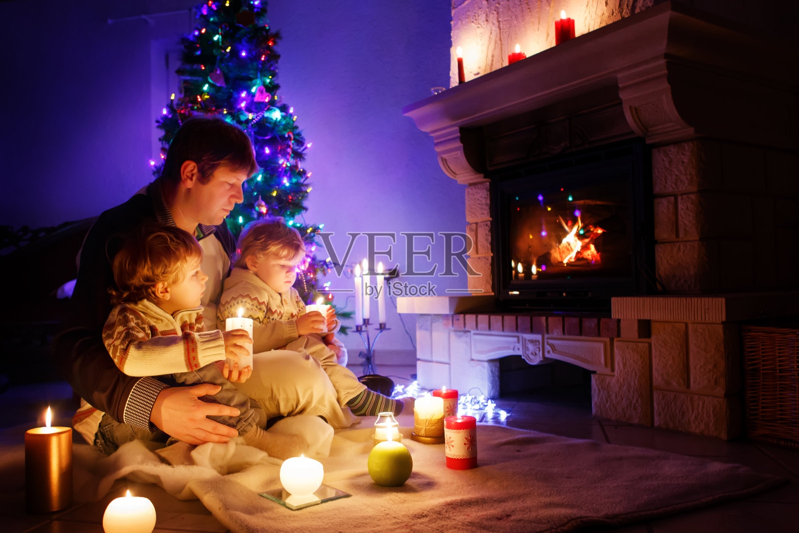 父亲和两个蹒跚学步的小男孩坐在烟囱、蜡烛和壁炉旁，看着火苗。家庭庆祝圣诞节。背景是圣诞树和彩灯。孩子们喜欢礼物照片摄影图片