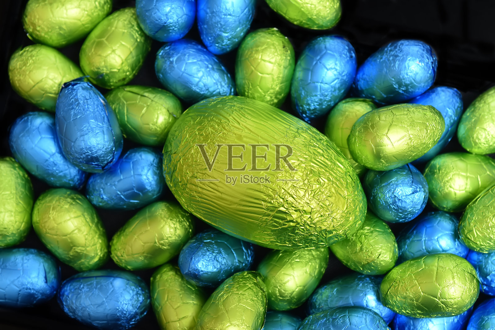 1 .用锡箔纸包裹一堆或一组颜色各异、大小各异的巧克力复活节彩蛋，颜色有蓝色、黄色和酸橙绿色，中间放一个绿色的大鸡蛋。照片摄影图片