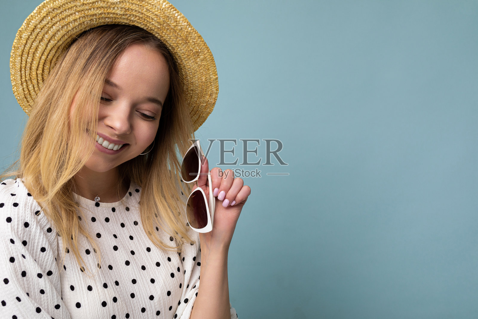 特写镜头相当积极的微笑年轻的金发女人戴着夏装草帽和时尚的太阳镜孤立在蓝色背景墙向下看照片摄影图片