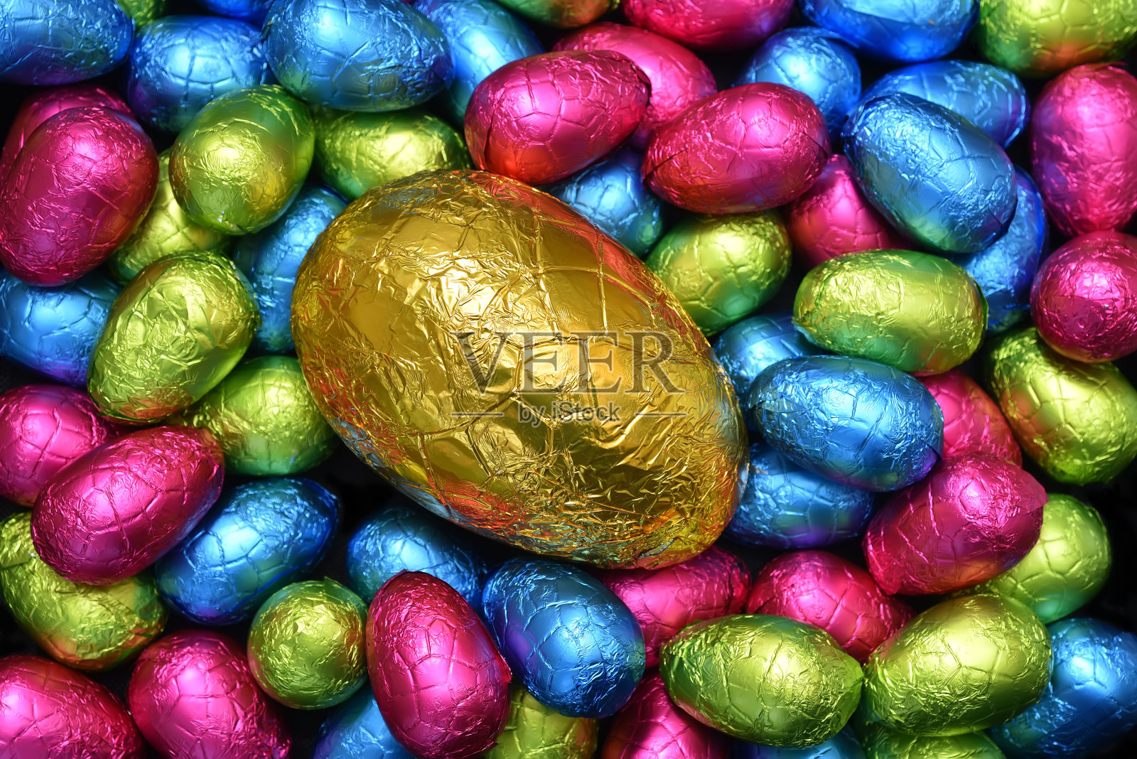 1 .用锡箔纸包裹一堆或一组颜色各异、大小不同的巧克力复活节彩蛋，彩蛋的颜色有粉色、蓝色、黄色和酸橙绿色，中间放一个大的金蛋。照片摄影图片