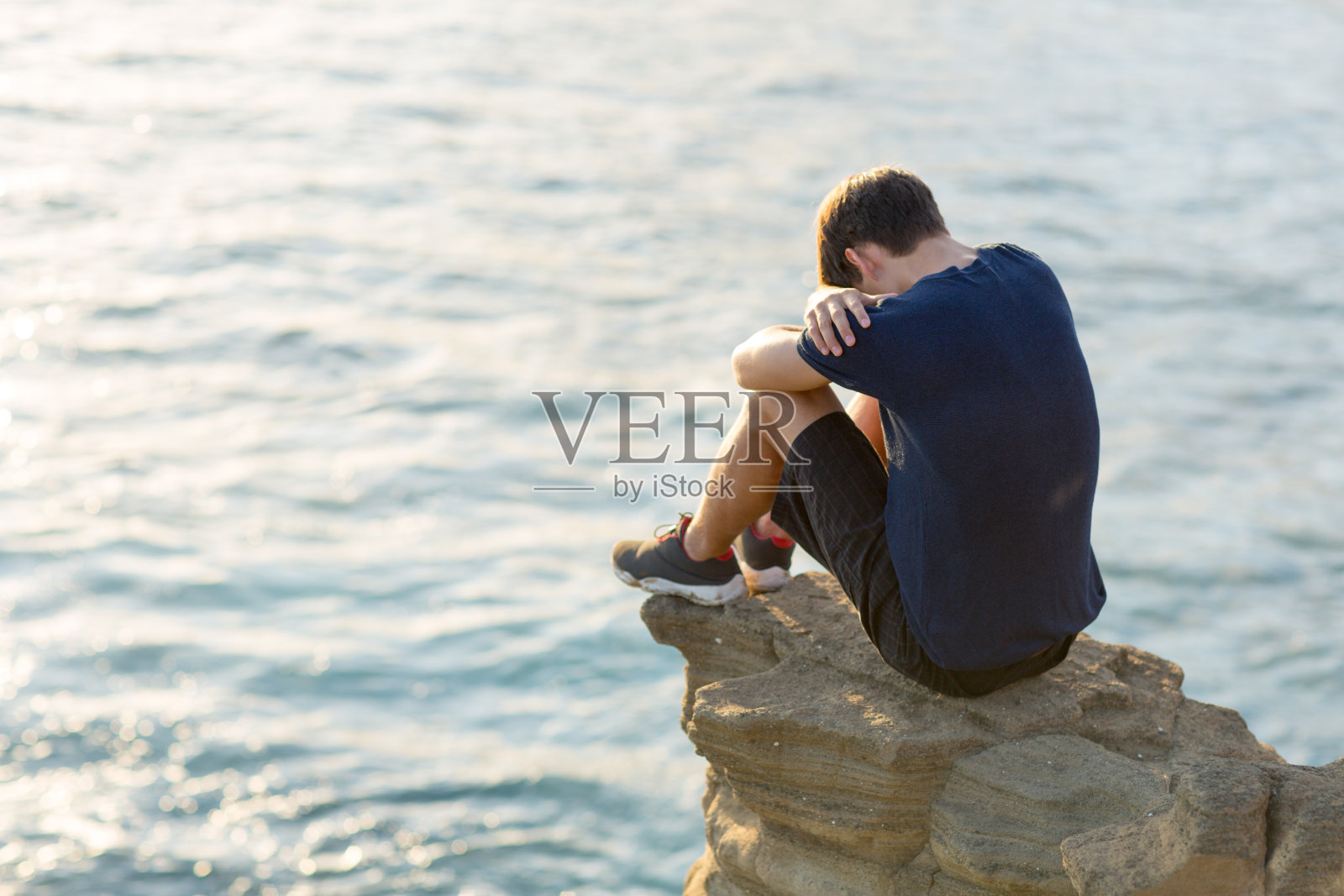 一个悲伤的年轻人独自坐在海边的悬崖上。照片摄影图片