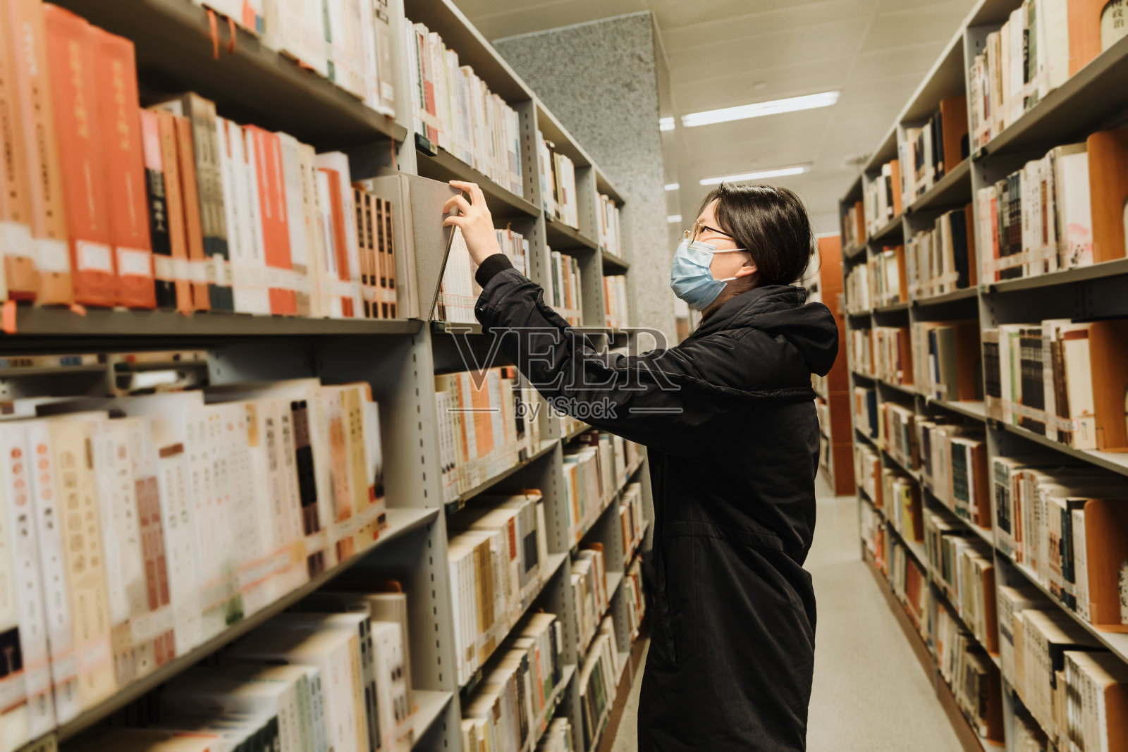 年轻的亚洲妇女戴着保护面罩在图书馆的书架上选择书籍照片摄影图片