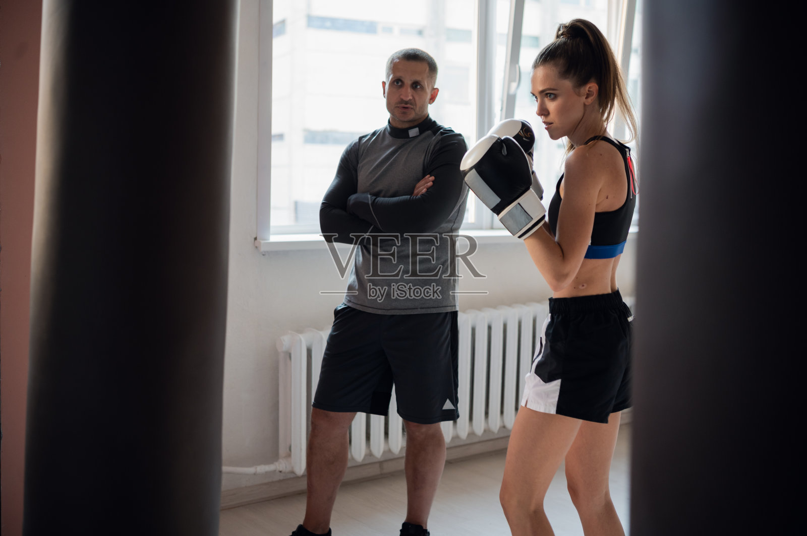 在一个宽敞的训练室里，一个严格的教练看着他的女自由搏击手学生在一个沙袋上练习出拳照片摄影图片